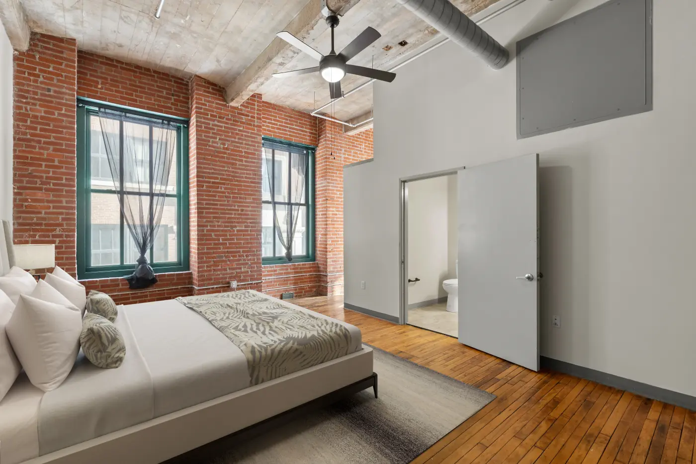 Bedroom with exposed brick, tall green-framed windows, a ceiling fan and an adjoining bathroom